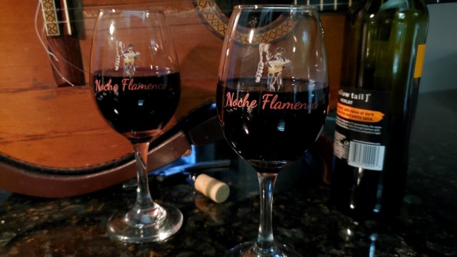 Two Noche Flamenca wine glasses, half full of red wine, stand in front of a guitar laying on its side.